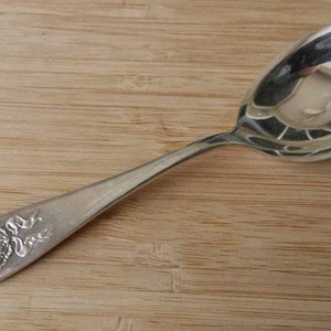 Puede incluir: Una cuchara de sopa de plata con un diseño floral en el mango. La cuchara está sobre una superficie de madera.