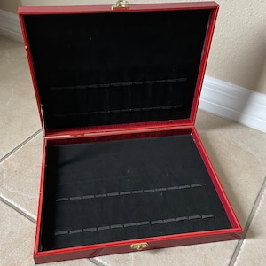 Puede incluir: Una caja de madera roja abierta con un interior de terciopelo negro. La caja tiene un cierre dorado y está diseñada para contener varios artículos.