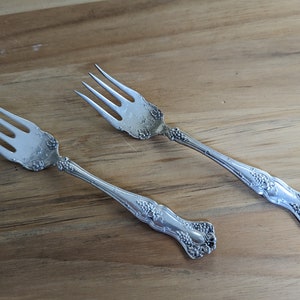 Può includere: Due forchette d'argento con disegni decorativi, che presentano un motivo a vite, sono esposte su una superficie di legno.