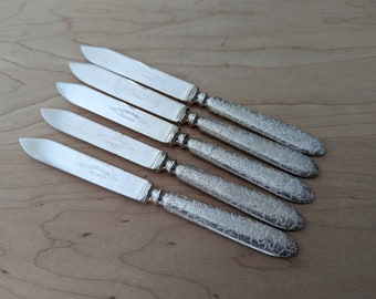 May include: Six silver butter knives arranged diagonally on a light wood surface. The blades are smooth and rectangular, with a slight curve at the tip. The handles feature an ornate, floral pattern. The knives are likely vintage or antique.