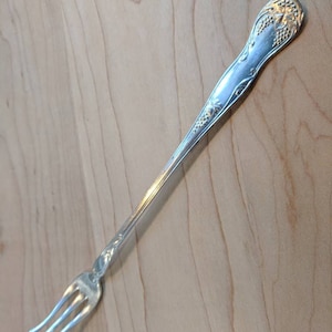 May include: A silver fork with an ornate handle featuring a grapevine design. The fork has three tines and is approximately 7 inches long.