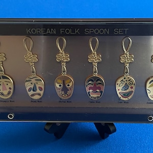 May include: A set of six Korean folk spoon keychains, each featuring a different traditional mask design. The spoons are gold-colored and have a knotted string attached. The masks are colorful and depict various expressions. The set is displayed in a black frame with a clear plastic cover. The text "KOREAN FOLK SPOON SET" is printed on the frame.