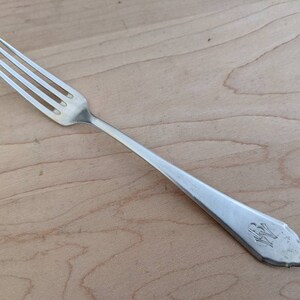 Puede incluir: Un tenedor de plata con un mango decorativo. El mango tiene un diseño floral y las iniciales "WB" grabadas en él.
