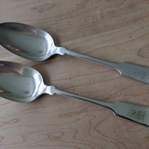 May include: Two antique silver spoons with a reflective surface, resting on a wooden surface. Each spoon has a long handle and a rounded bowl. The handles are engraved with an ornate monogram.