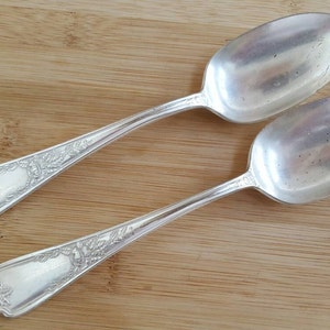 May include: Two silver spoons with ornate handles. The spoons are lying on a wooden surface.