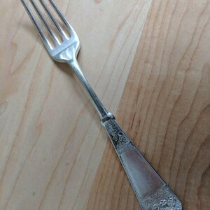 May include: A silver fork with an ornate handle. The handle has a floral design and a raised border.