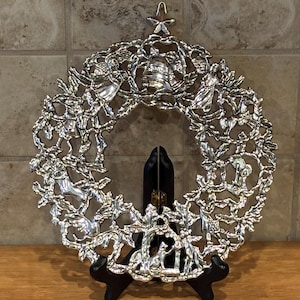 May include: Silver metal Christmas wreath with detailed depictions of angels, bells, and deer. The wreath is displayed on a black stand, showcasing its intricate design and festive theme. A star sits at the top.