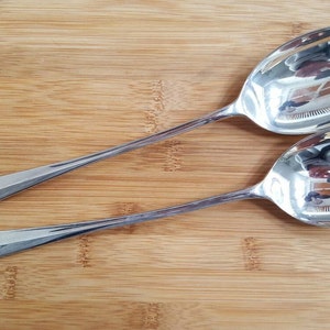 May include: A silver salad serving set with a large spoon and a fork with three tines. The set is on a wooden surface.