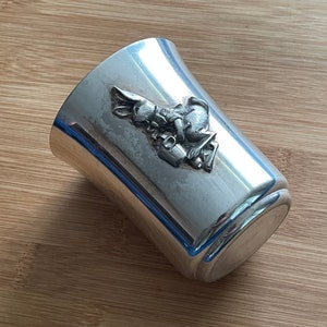 May include: Silver cup with a rabbit design. The rabbit is holding a bucket and is sitting on a flower.