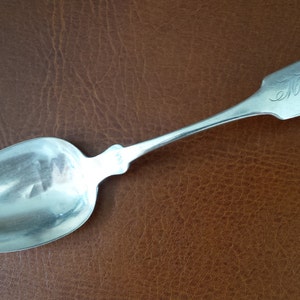 Può includere: Un cucchiaio d'argento con una ciotola arrotondata e un manico lungo e sottile. Il manico è inciso con il testo "E.M. Barnard".