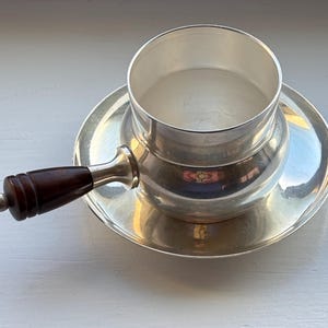 Op de afbeelding: Een kleine, zilverkleurige steelpan met een donkerbruine houten handgreep en een bijpassende schotel. De handgreep is aan de zijkant van de steelpan bevestigd met een zilveren metalen fitting. De set is reflecterend en staat op een wit oppervlak.