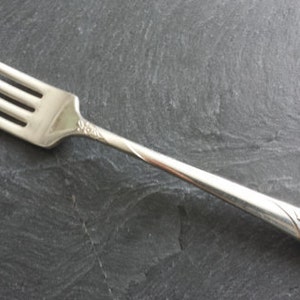 Puede incluir: Un tenedor de metal plateado con cuatro dientes y un mango delgado. El mango está decorado con un patrón floral en relieve en la base y un detalle floral más pequeño donde el mango se une con el cuello del tenedor.