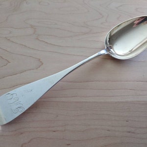 Puede incluir: Una cuchara de plata con un cuenco redondeado y un mango largo y delgado. El mango está grabado con el nombre "Sylvia".