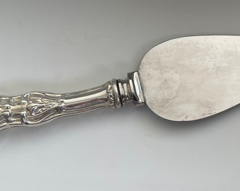 May include: A silver cheese knife with an ornate handle. The blade is smooth and slightly curved.