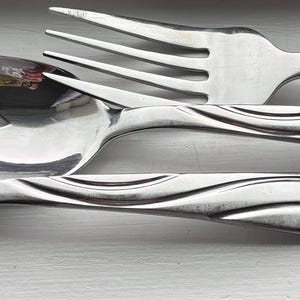 Puede incluir: Un juego de tres utensilios de acero inoxidable que consta de un tenedor, una cuchara ranurada y una cuchara sólida. Cada pieza presenta un acabado pulido y reflectante con un mango decorado con una serie de líneas onduladas paralelas en relieve.