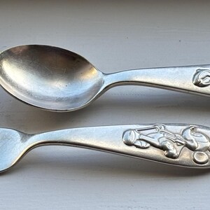 May include: A silver spoon and fork set. The handles feature a raised design of a cartoon character. The spoon has a rounded bowl, and the fork has three tines. The set is made of a shiny metal.