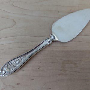 May include: A silver cheese knife with an ornate handle. The handle features a floral design and a raised crest.