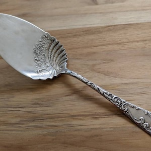 Puede incluir: Un servidor de pastel de plata con un diseño floral ornamentado en el mango y la hoja. El servidor está sobre una superficie de madera.