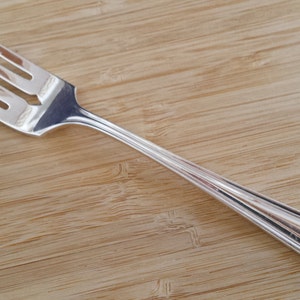 Puede incluir: Un tenedor de plata con un diseño sencillo y un mango ligeramente curvado. El tenedor está sobre una superficie de madera.