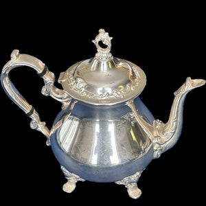 May include: A silver teapot with intricate detailing. The teapot has a curved spout, a decorative handle, and a lid with a finial. It stands on three small feet and has a reflective surface.