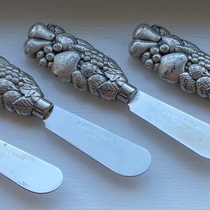May include: Four silver butter knives with detailed fruit and leaf designs on the handles. The knives have a polished finish and are ideal for spreading butter or soft cheese.