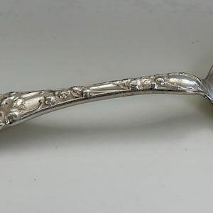 Puede incluir: Una cucharita de plata con un mango adornado y un monograma "A".