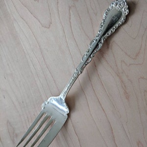 Könnte beinhalten: Eine silberne Torten Gabel mit einem verzierten Griff und einem dreizackigen Kopf. Der Griff hat ein dekoratives Design und ist mit dem Buchstaben "K" graviert.