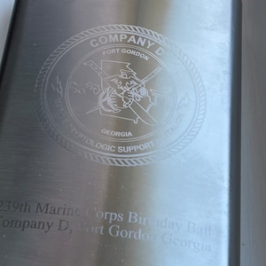 May include: A silver metal flask with an engraved inscription that reads "Company D, Fort Gordon, Georgia, 239th Marine Corps Birthday Ball". The flask has a circular emblem with the words "Marine Cryptologic Support Battalion, Georgia" and a stylized image of a state of Georgia.