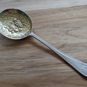 Pode incluir: Uma colher de prata com uma tigela folheada a ouro que apresenta um desenho gravado de frutas e folhas. O cabo é de prata lisa com um adorno decorativo na extremidade.