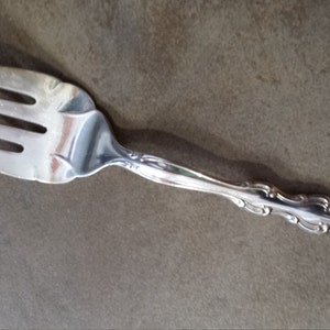 Può includere: Una forchetta d'argento con quattro rebbi e un manico decorativo. La forchetta è in metallo lucido e si trova su una superficie marrone. Il manico presenta un ornato motivo floreale.