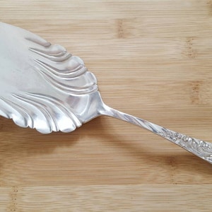 Puede incluir: Un servidor de pastel de plata con un borde festoneado ornamentado y un mango retorcido. El mango está decorado con un diseño floral.