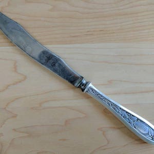 May include: A silver butter knife with an ornate handle. The handle has a floral design and a small oval with an inscription.