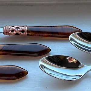 May include: A collection of eight spoons with shiny silver bowls and dark brown handles. The handles are accented with a decorative copper-colored band. The spoons are arranged on a light-colored surface, suggesting a dining or serving context.