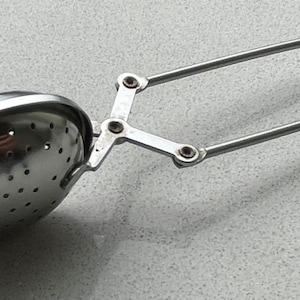 May include: A silver tea infuser with a hinged handle and a round, perforated basket. The infuser is designed to hold loose tea leaves and steep them in hot water.