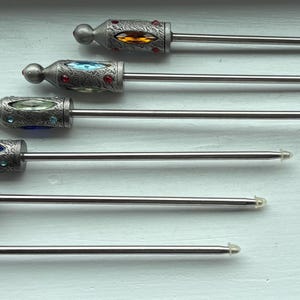 May include: A collection of six ornate silver-toned bottle stoppers. Each stopper features a decorative top with a jewel-toned gem in red, orange, blue, green, and yellow. The stoppers have a long, slender, silver-toned stem.