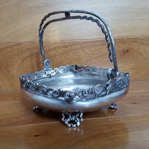 Può includere: Un cesto da portata in argento con un manico decorativo e dettagli decorativi. Il cesto ha una forma quadrata con un bordo a festoni e poggia su quattro piedini.