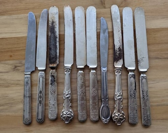 May include: A set of ten silver butter knives with ornate handles. The knives have varying degrees of wear and tarnish.
