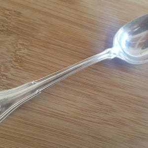 May include: A silver teaspoon with a rounded bowl and a decorative handle. The handle has a raised design and a small, engraved mark.