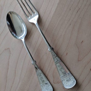 May include: A silver fork and spoon with ornate designs and engraved lettering. The fork has four tines and the spoon has a rounded bowl.