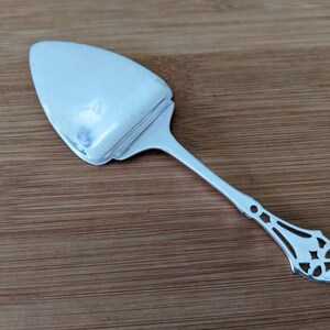 Puede incluir: Un servidor de pastel de plata con un mango adornado y recortado. El servidor tiene una hoja ancha y plana y un mango largo y delgado.