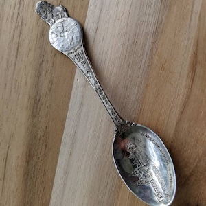 Puede incluir: Cuchara de recuerdo de plata con un león y un globo en el mango y una imagen de un edificio en el cuenco. La cuchara está grabada con "Alrededor del mundo en 1901" y "Ciudad de Nueva York".