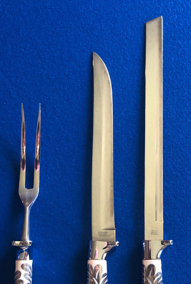 May include: A set of three silver-toned kitchen utensils with ivory-colored handles. The set includes a carving knife, a bread knife, and a fork. The handles have a floral design.
