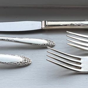 Könnte beinhalten: Ein Satz antiker Silberbestecke, darunter zwei Löffel, ein Messer und zwei Gabeln. Das Besteck ist silberfarben mit aufwendigen floralen Details an den Griffen. Die Utensilien sind auf einer hellgrauen Oberfläche angeordnet.