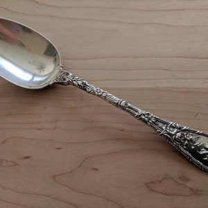 Puede incluir: Una cuchara de plata con un diseño floral ornamentado en el mango. La cuchara está pulida y brillante.