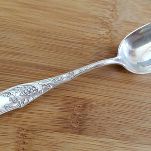 Puede incluir: Una cuchara de plata con un mango adornado y grabado. La cuchara tiene un cuenco redondeado y un mango largo y delgado.