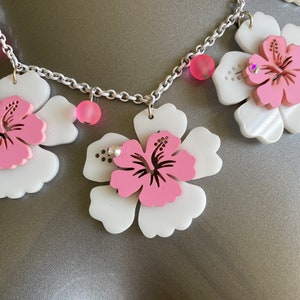 May include: A necklace featuring large white and pink hibiscus flower pendants. The flowers have a pink center and are accented with small pink beads and rhinestones. The chain is silver-colored.