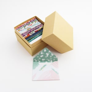 May include: A kraft paper box with a lid, open to reveal a collection of colorful stationery. An open envelope with a green polka dot interior and pink and green abstract design sits in front.