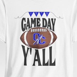 May include: White t-shirt with a football graphic and the text "IT'S GAME DAY Y'ALL". The football is brown with white stripes and a blue logo. Blue pennants are above the words "GAME DAY".