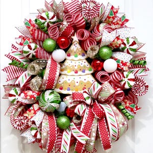 Puede incluir: Una corona navide&ntilde;a festiva con cintas, adornos y un centro de mesa de &aacute;rbol de pan de jengibre rojo, blanco y verde. La corona presenta varios patrones, incluyendo rayas, lunares y dise&ntilde;os tem&aacute;ticos de vacaciones, creando una exhibici&oacute;n alegre y decorativa.