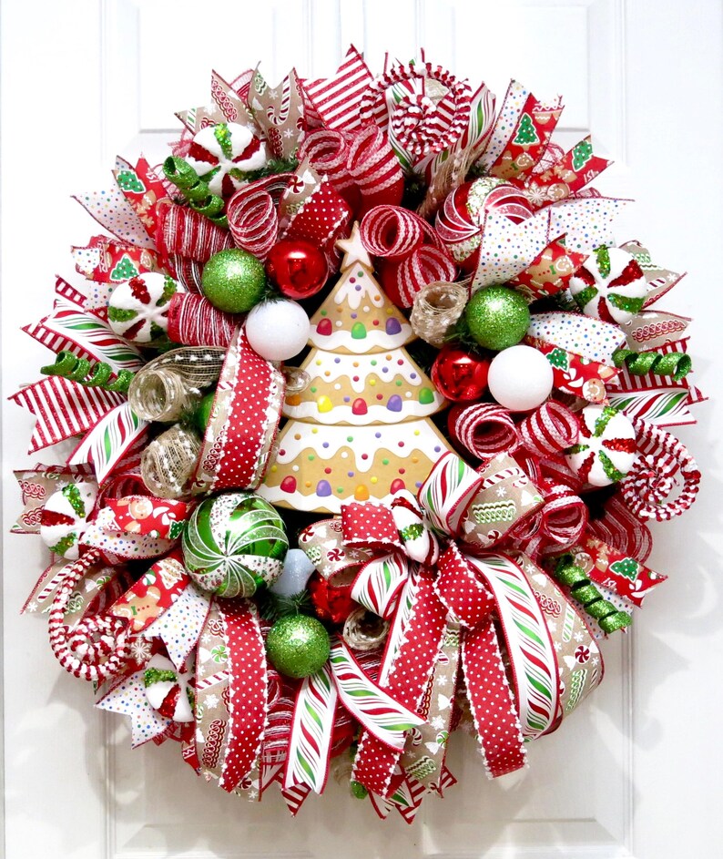 Puede incluir: Una corona navide&ntilde;a festiva con cintas, adornos y un centro de mesa de &aacute;rbol de pan de jengibre rojo, blanco y verde. La corona presenta varios patrones, incluyendo rayas, lunares y dise&ntilde;os tem&aacute;ticos de vacaciones, creando una exhibici&oacute;n alegre y decorativa.